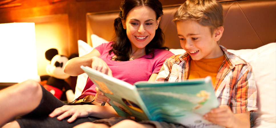 In the comfort of the Disneyland Hotel room, a boy reads a Disney storybook to his mom.