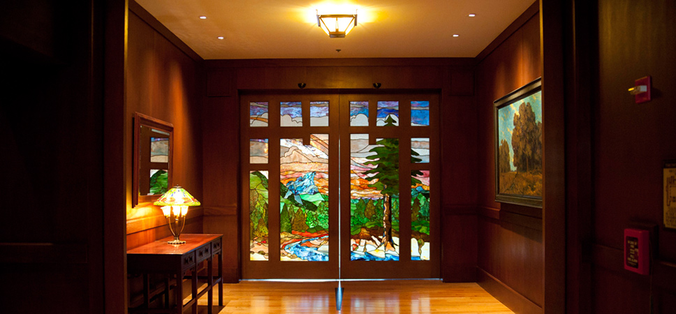 Stained glass doors at the end of the hallway show a scene with a bright blue sky, lush evergreen trees and a sparkling creek.